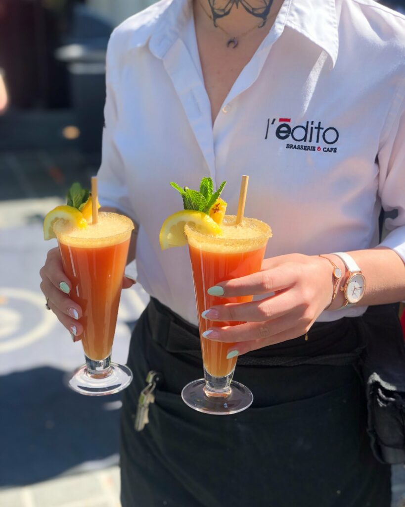 Serveuse de L’Édito portant deux cocktails orange garnis de menthe et d’ananas.